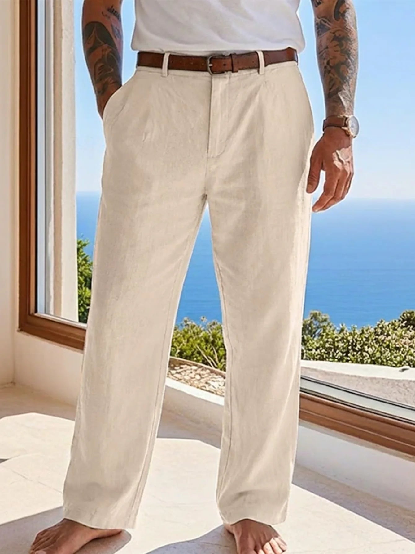A man wearing off-white linen trousers with a brown leather belt, standing barefoot on a sunlit balcony with a blue ocean view in the background.