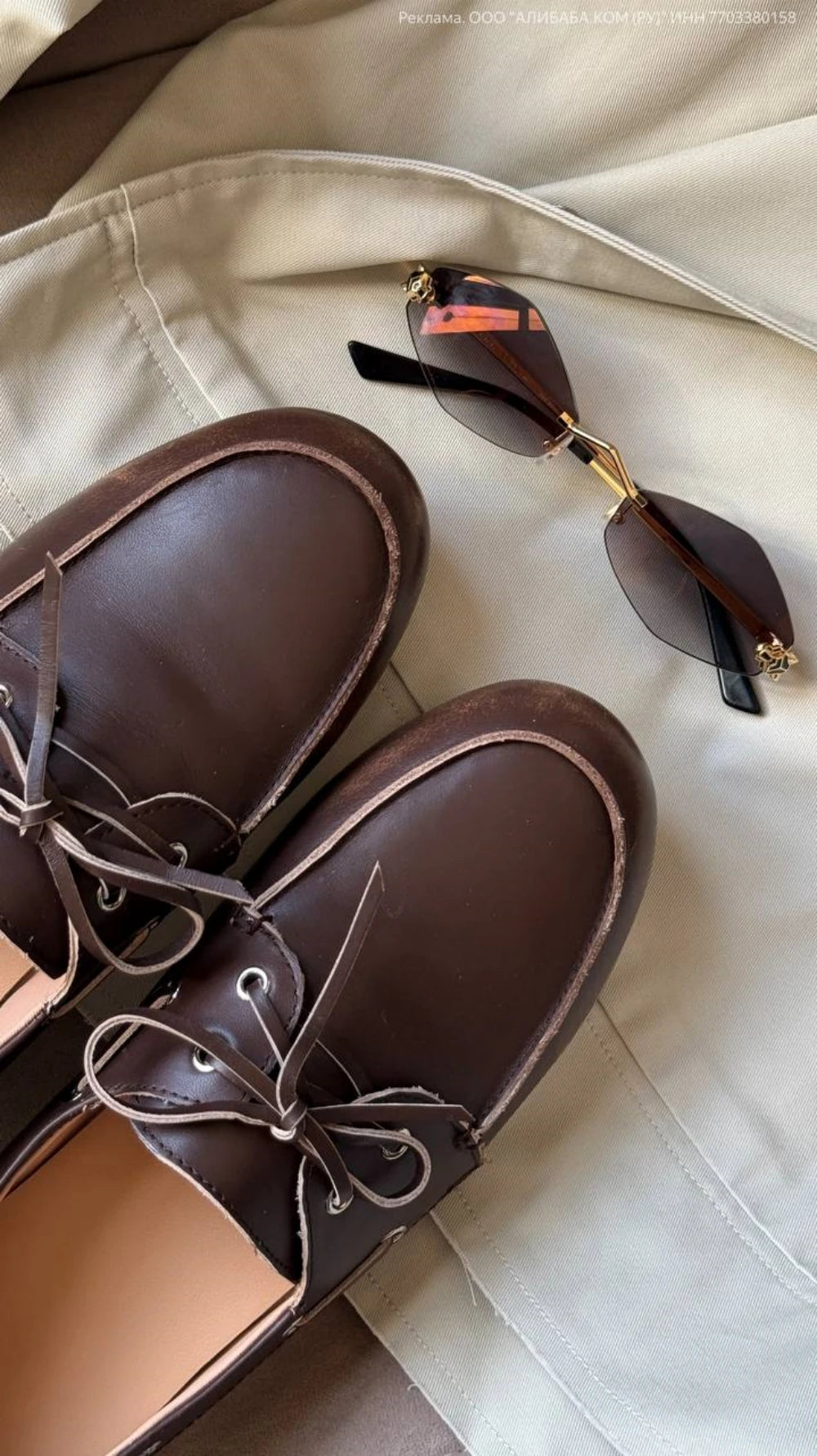 A pair of dark brown leather boat shoes with matching laces, styled on a beige fabric background next to a pair of sunglasses.