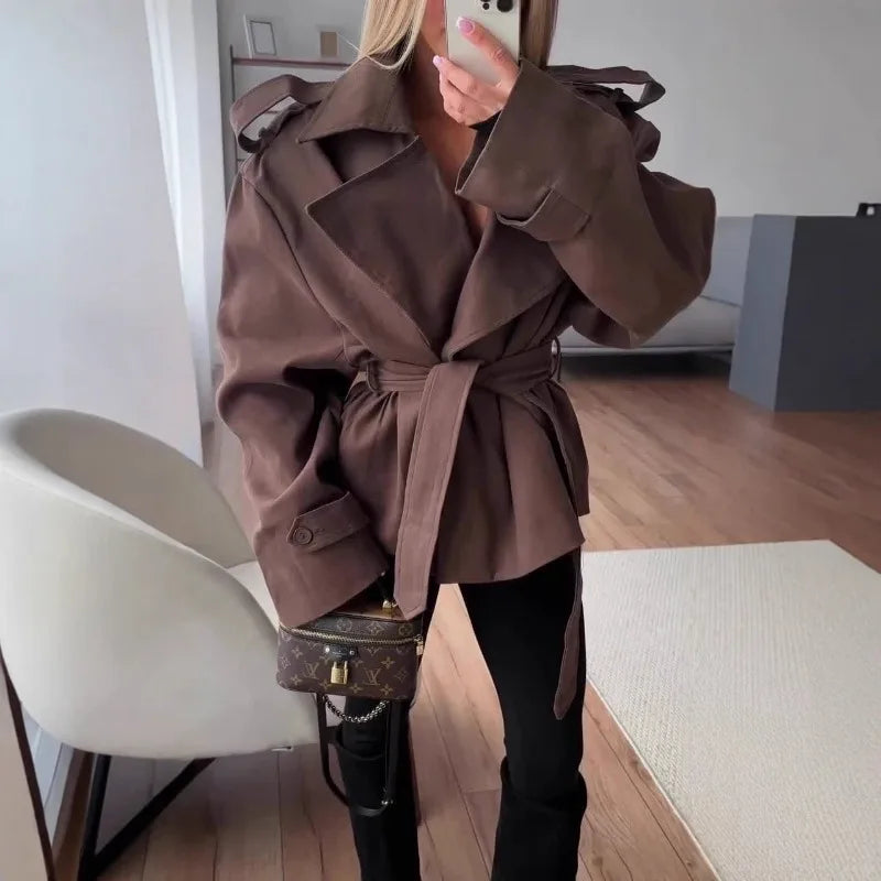 Person wearing a brown trench coat taking a mirror selfie indoors.