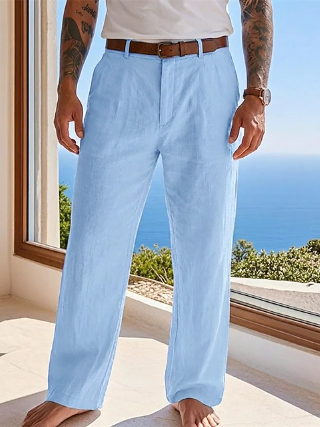 A man wearing light blue linen trousers with a brown belt, standing barefoot on a sunlit balcony with a view of the blue ocean in the background.