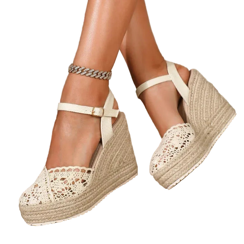 A pair of women's espadrille wedge sandals with cream-colored lace-up straps and openwork lace detailing on the toe, shown on a model's feet. The sandals feature braided jute wedge heels and are set against a neutral indoor background with a textured wall and floor.