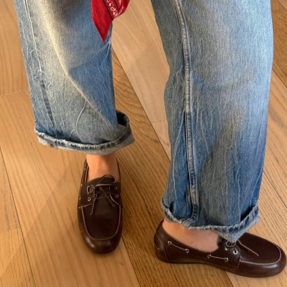 Brown leather shoes worn with rolled-up blue jeans on a wooden floor.